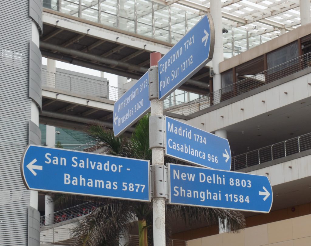 Zu sehen ein Schild was in 5 Himmelrichtung zeigt. Die oberen zwei Schilder zeigen Richtung Norden und Süden. Zu lesen Amsterdam 3173 Km. Bruslas 3020 Km.  Sapetown 7741 Km. Richtung ost New Delhi 8803 Km. Shanghai. Richtung West, San Salvador- Bahamas 5877 Km. Das letzte Schild Richtung Richtung Nord Ost zu lesen Madrid 1734 Km. Casablanca 966 Km.